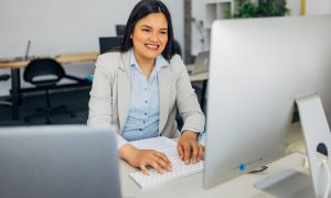 Woman typing on a keyboard in an office setting, with a large monitor in front of her.