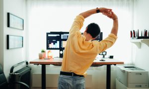 Man standing and stretching in front of a desk with monitors displaying charts.