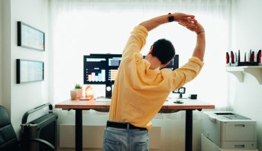 Man standing and stretching in front of a desk with monitors displaying charts.