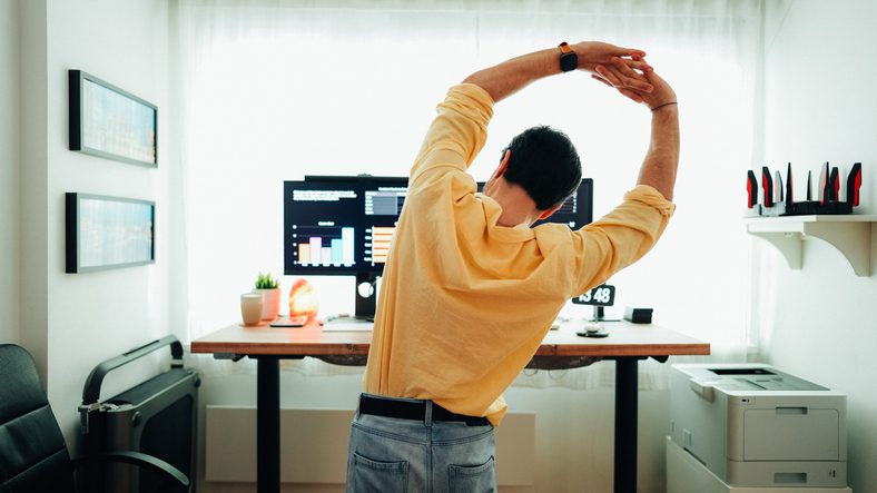 Man standing and stretching in front of a desk with monitors displaying charts.