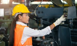 Woman in a hard hat, safety vest, and gloves operating machinery in a factory.