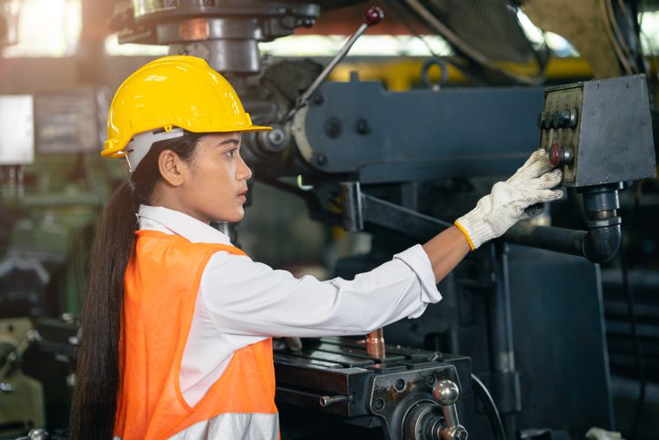 Woman wearing a hard hat, safety vest, and gloves operating machinery in a factory.