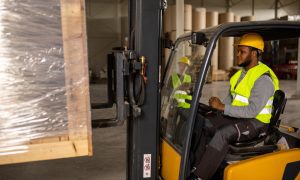 A worker drives a forklift in a warehouse, lifting a wrapped pallet.