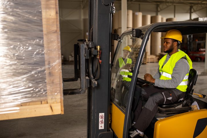 A worker drives a forklift in a warehouse, lifting a wrapped pallet.