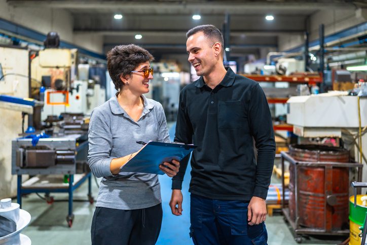 Two individuals are standing in a factory or industrial setting. One person is holding a clipboard and appears to be discussing something with the other person. The background shows various machinery and equipment typically found in a manufacturing environment. Both individuals have their faces obscured for privacy.