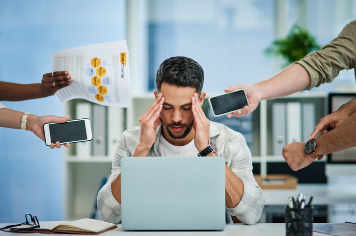 Stressed man at a desk with a laptop, holding their head while multiple hands reach in with phones and documents in an office setting.