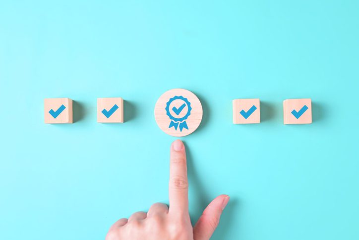 A hand points at a wooden block with a ribbon and checkmark, surrounded by similar blocks.