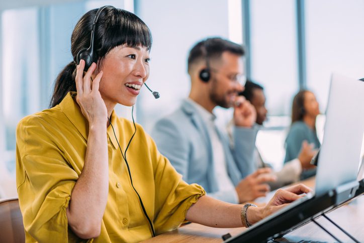 Customer service team in modern office using headsets and laptops for support calls, with natural light from large windows enhancing productivity.