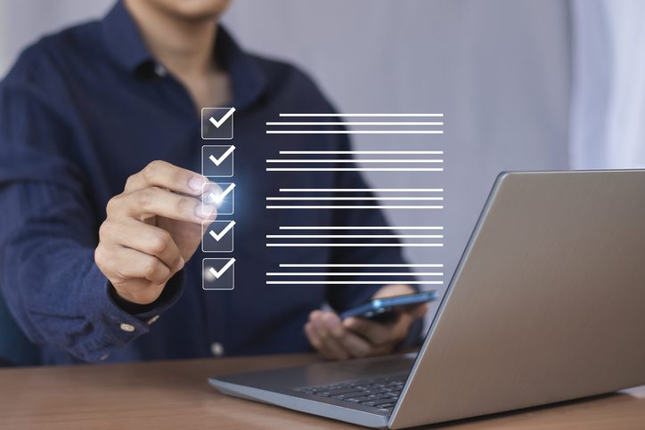 Person in a dark blue shirt using a laptop and holding a smartphone, interacting with a virtual checklist featuring five items marked with checkmarks.