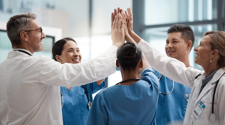 Group of healthcare professionals in medical uniforms and lab coats standing together in a hospital setting, raising their hands for a high-five in a gesture of teamwork and celebration.