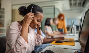 Black woman wearing a headset and working at a computer in a modern office setting, with other team members collaborating in the background.