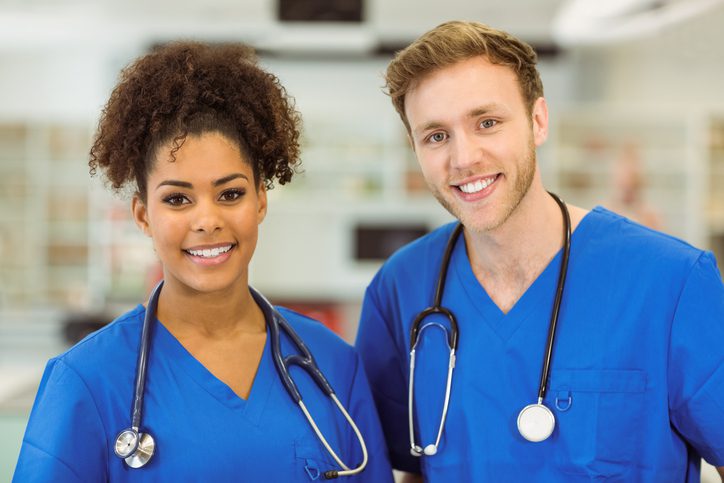 Two healthcare professionals wearing blue medical scrubs and stethoscopes stand side by side in a brightly lit hospital setting, smiling warmly at the camera. The background features blurred medical equipment, emphasizing a clinical environment and a sense of teamwork and professionalism.