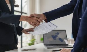 Two people in business attire shaking hands across a desk, one holding a document labeled ‘Resume.’ A laptop displaying a chart and a pen are visible on the desk.