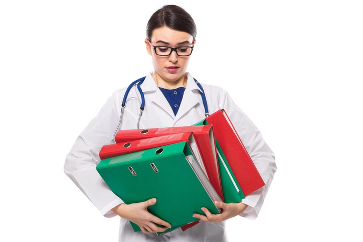 A medical professional in a white lab coat with a stethoscope around their neck struggles to carry a large stack of colorful binders, conveying a sense of being overwhelmed by paperwork. The background is plain white, emphasizing the subject and the burden.