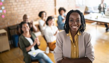 Photo of a Black woman standing in the foreground with arms crossed, wearing a light-colored blazer over a yellow shirt. In the background, several people are seated in a meeting, with a brick wall and sticky notes visible. A table with papers and a laptop is positioned near a window, suggesting a collaborative workspace.