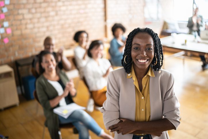 Photo of a Black woman standing in the foreground with arms crossed, wearing a light-colored blazer over a yellow shirt. In the background, several people are seated in a meeting, with a brick wall and sticky notes visible. A table with papers and a laptop is positioned near a window, suggesting a collaborative workspace.