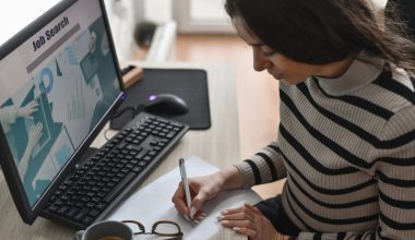 Person writing on paper at a desk with a computer screen showing “Job Search” and charts; keyboard, mouse, coffee cup, and eyeglasses nearby.