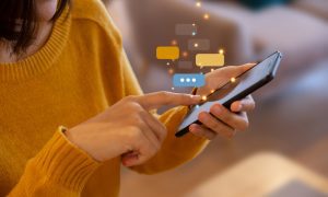 Photo of a woman with medium‑length dark hair wearing a mustard yellow sweater, using a smartphone indoors. She taps the screen with one hand while holding the phone in the other. Colorful illustrated chat bubbles float above the phone, symbolizing messaging or social interaction.