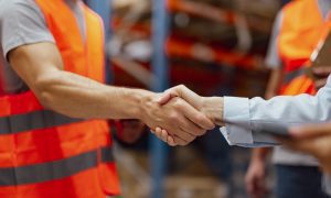 Photo of two people shaking hands in a warehouse. One man wears an orange safety vest and gray shirt; the other person wears a light blue long‑sleeve shirt. Shelving and another person in an orange vest are visible in the background.