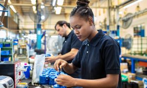 Photo of two assembly line workers in a factory setting; a foreground worker appearing female with hair in a braided bun wears a black short‑sleeve polo shirt and uses a hand tool to assemble blue mechanical components on a workbench, while a background worker appearing male in a black shirt performs a similar task along the same production line.