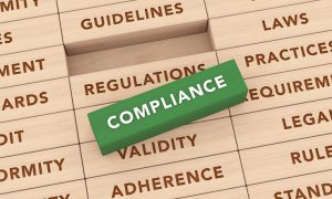 Photo of a green rectangular block labeled “COMPLIANCE” placed diagonally on stacked wooden blocks engraved with words including “REGULATIONS,” “GUIDELINES,” “LAWS,” “REQUIREMENTS,” “LEGAL,” “RULES,” “STANDARDS,” “VALIDITY,” “ADHERENCE,” “CONFORMITY,” and “AUDIT.”