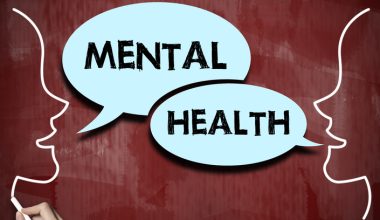 Photo illustration of two outlined human profiles facing each other on a red chalkboard background, with blue speech bubbles reading “MENTAL HEALTH.” A hand holding white chalk appears in the lower-left corner.