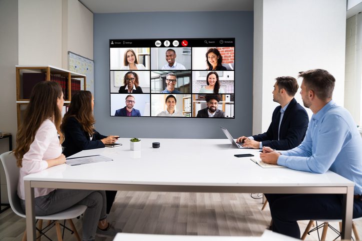 Five people meet in a conference room while a video call with nine remote participants plays on a large screen.