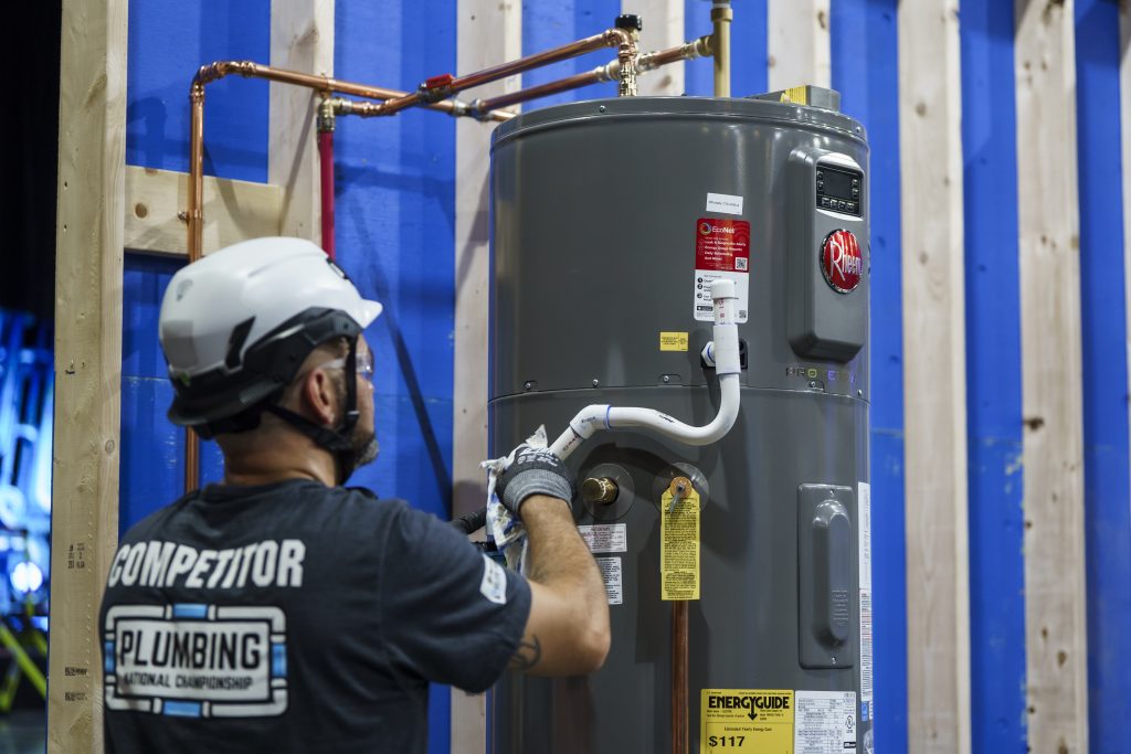 Photo of an adult man wearing a white safety helmet, gray gloves, and a black T‑shirt with the text “COMPETITOR” and “PLUMBING National Championship” on the back, installing a gray water heater connected to copper piping in an indoor construction setting. 