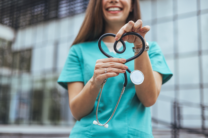 A healthcare worker in teal scrubs holds a stethoscope shaped into a heart, standing in front of a modern glass building.