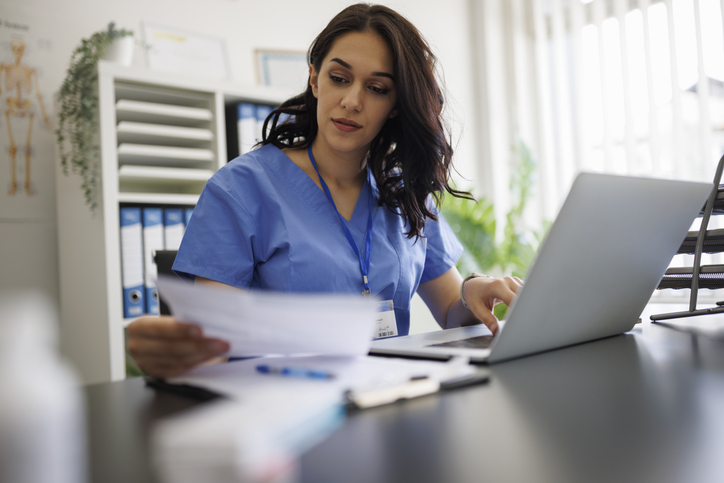 Focused female healthcare leader in blue scrubs reviews paperwork and types on a laptop, illustrating aligned healthcare operations and financial management.