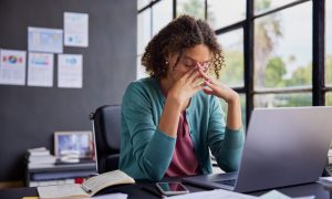 Photo: adult woman with curly hair wearing a green cardigan over a pink top sits at a desk, eyes closed and fingers pressed to her face, appearing stressed while working on a laptop with papers, notebook, and phone nearby.