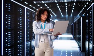 Photo: adult woman with curly dark hair wearing a light blue shirt, ID lanyard, and beige pants, using a laptop while standing in a data center aisle lined with illuminated server racks.