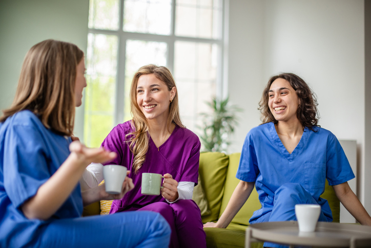 Three young female nurses are enjoying a break together, chatting and laughing while holding cups. They are dressed in colorful scrubs in a bright healthcare setting, discussing flexible schedules.