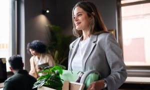 Photo of a person in a light gray blazer and white shirt carrying a box with a green plant and office supplies in a modern office; two other people are blurred in the background near desks and windows.